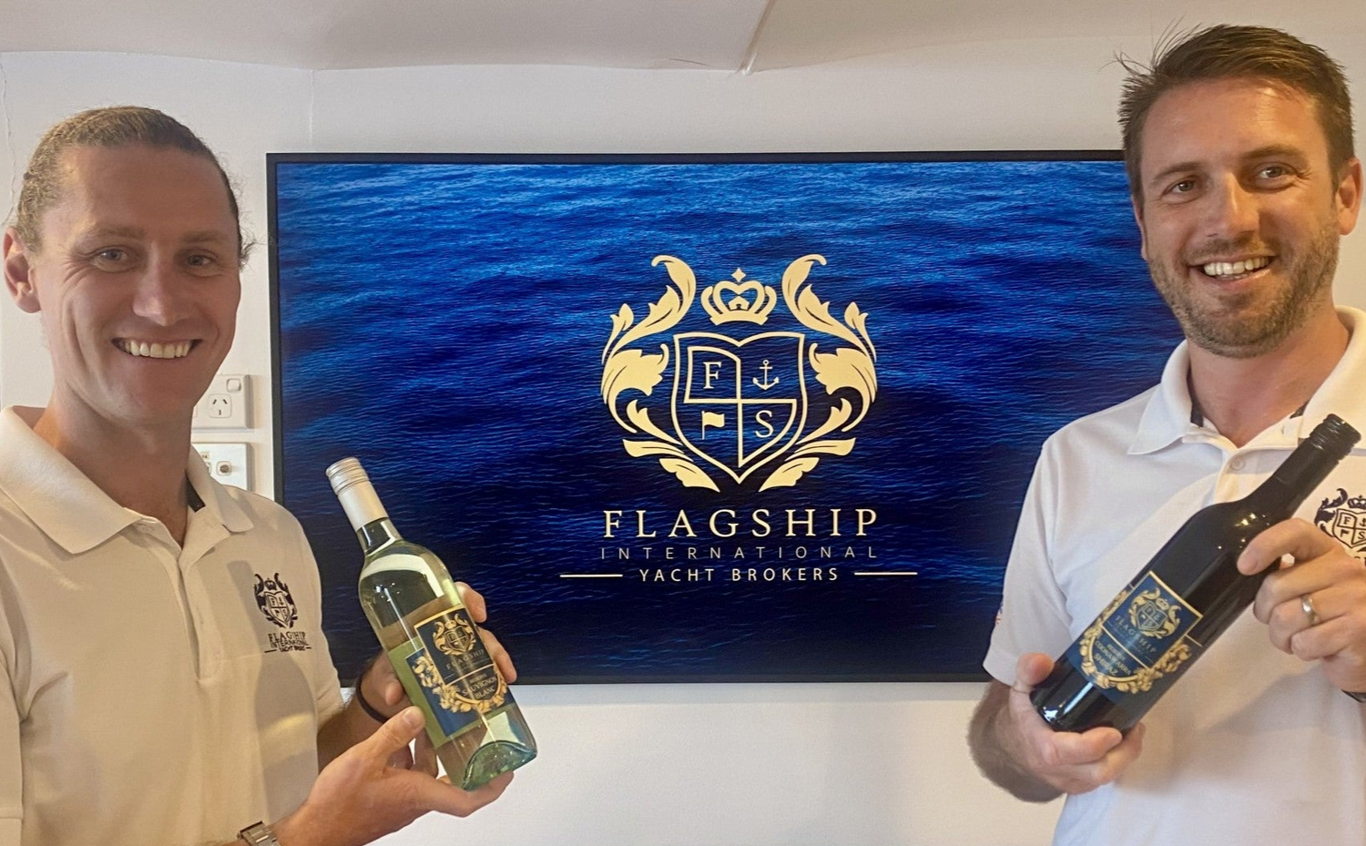 Two men holding bottles of alcohol in front of a Flagship Yachts logo.